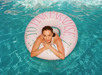 Woman floating on a pink inflatable ring with 'Eden Paraiso' text in clear blue water.