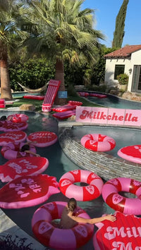Pool with heart-shaped inflatable rings and 'Milchella' branding, surrounded by palm trees and a house.