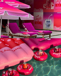 Pink lounge chairs and inflatable pool floats with poppi cans in a pool area.