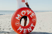 Person holding a red 'MOVEMENT' inflatable ring on a beach