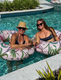 Two women enjoying a drink on inflatable pool floats in a pool.