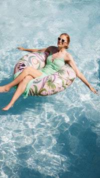 Woman in a green dress and sunglasses lounging on a floral pool float in clear blue water.